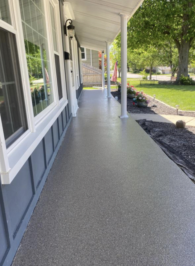 A residential front porch with gray flooring and white columns stretches alongside a house with white siding and windows, overlooking a landscaped front yard.