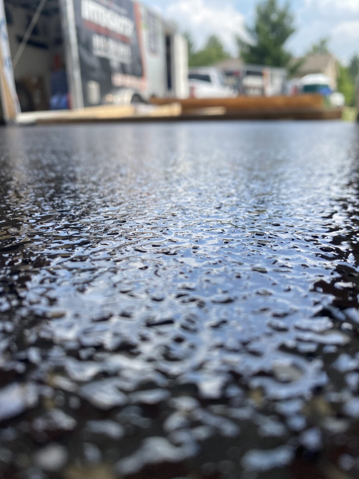 Close-up of a wet surface reflecting the sky, with water droplets scattered across. In the background, there's an unfocused view of vehicles and a trailer.