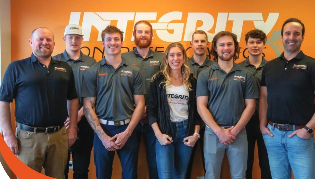 A group of nine people, wearing matching branded attire, are smiling and posing for a photo in front of an orange wall with the word "INTEGRITY."