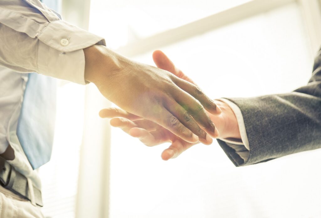 Two people are shaking hands, backlit by a bright window. One is wearing a gray suit; the other has a light blue shirt.