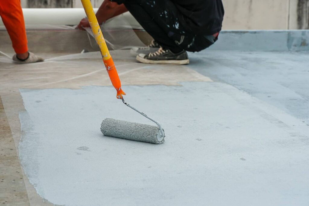 A person is applying a coating to a concrete surface using a paint roller with an extension pole. Construction or renovation work seems in progress.