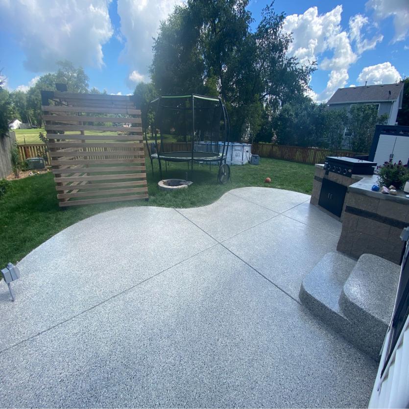 A sweeping, textured patio leads to a lush backyard with a wooden privacy screen, trampoline, and outdoor kitchen under a clear blue sky.